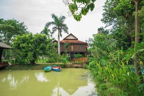 A traditional wooden house with a thatched roof, surrounded by lush greenery and a calm pond with boats. - Hok Sokol's Villa by Ream Lek (Krong Siem Reap)