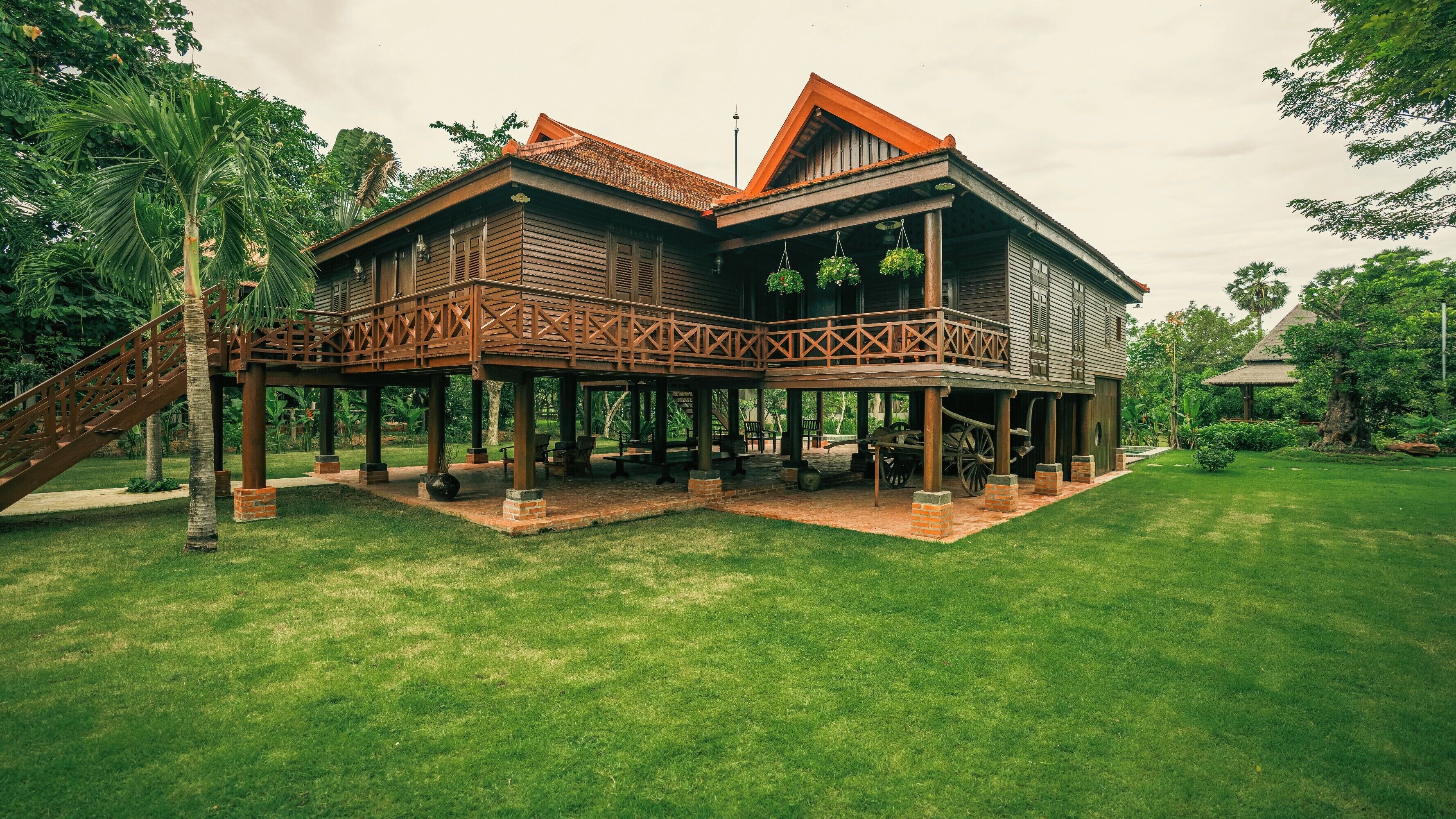 A two-story wooden house with a balcony, surrounded by a well-maintained lawn and tropical plants.