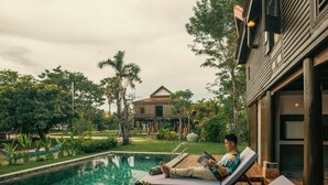 Un hombre descansando en una silla de salón junto a la piscina, con un cuenco de fruta en la mesa y una vista de una casa al fondo.