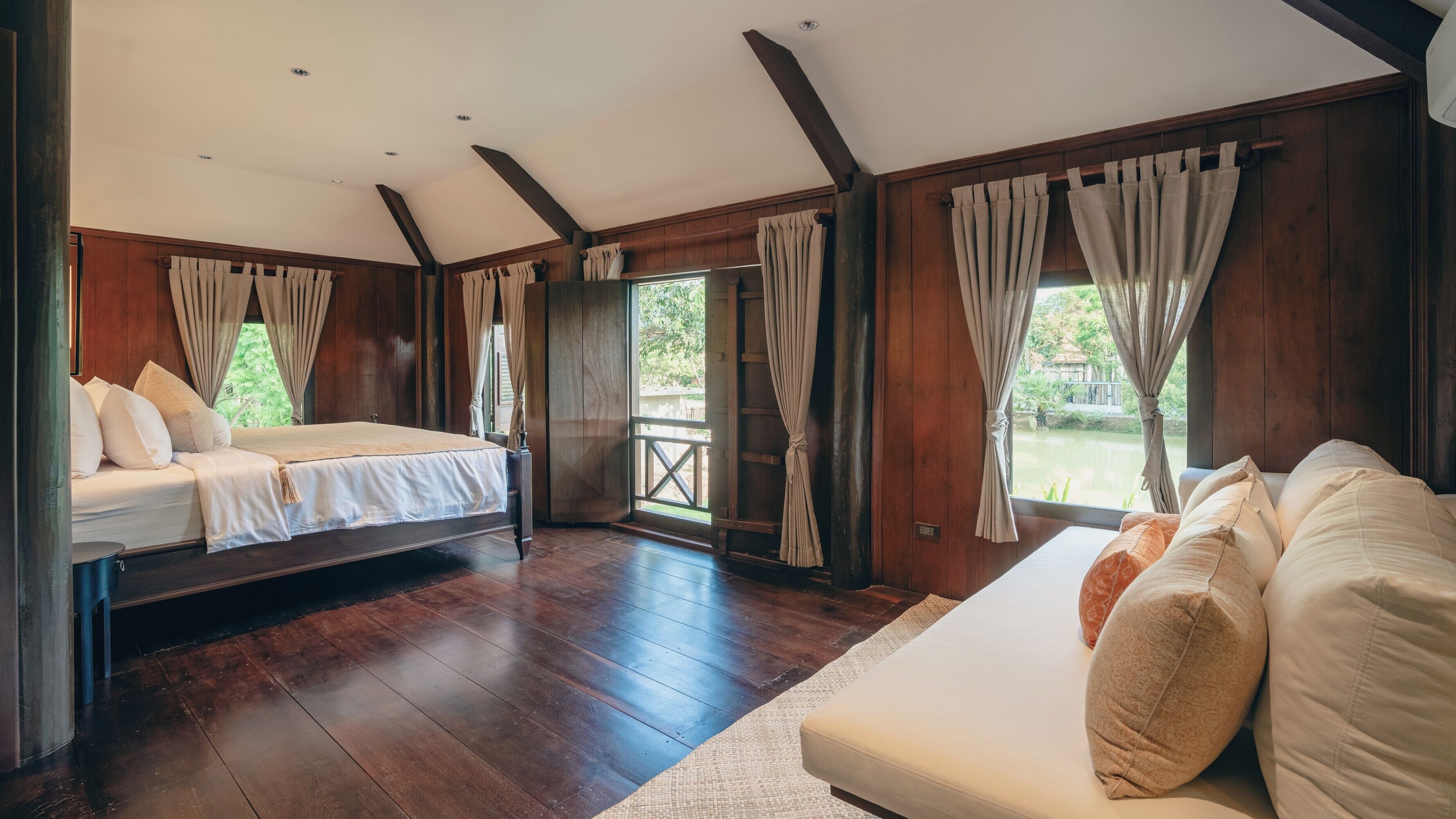 A spacious bedroom with a large bed, a sofa, wooden flooring, and a view of greenery outside.