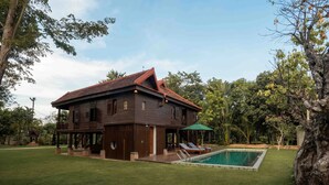 Una casa de madera de dos pisos con piscina, rodeada de césped cuidado y vegetación tropical.