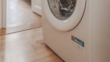 A front-loading washing machine in a modern kitchen with a wooden floor and a view of a dining area.