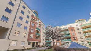 Edifício residencial com vários andares, varandas, um pequeno jardim e céu limpo.