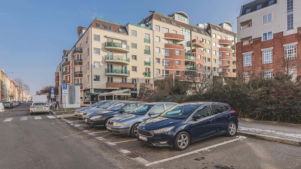 En flere-etagers boligbygning med altaner, en parkeringsplads med biler, og en klar himmel.