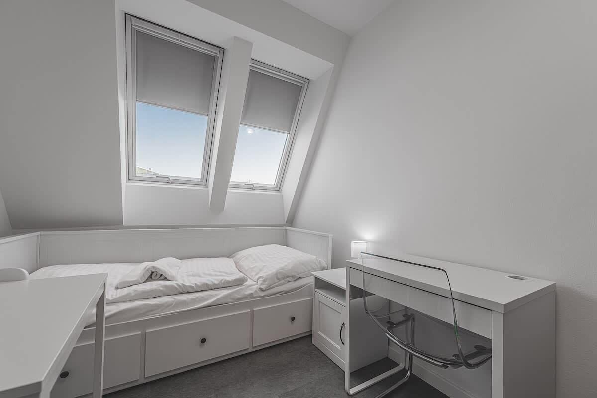 Una habitación pequeña y blanca tipo dormitorio con una cama litera, un escritorio y una ventana.