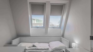 Pieni, valkoinen huone, jossa on yksi sänky, kalteva katto ja ikkuna, josta avautuu näkymä toiseen rakennukseen.