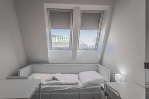 Ein kleines Zimmer mit einem Einzelbett, weißen Wänden und schrägem Dach. Ein Fenster bietet Blick auf ein Gebäude.