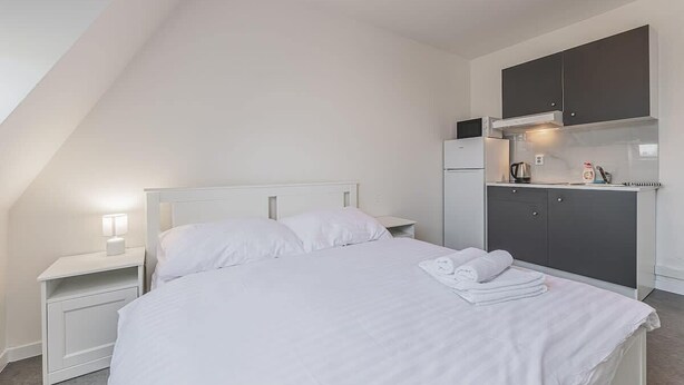 Um quarto com uma cama, mesas de cabeceira brancas, uma escrivaninha pequena e uma cozinha compacta.