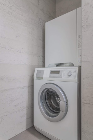 A front-loading washing machine in a corner of a room with a textured white wall. - Zizkov flat/skyline views/AC/Kitchen, new bathroom (Prague)