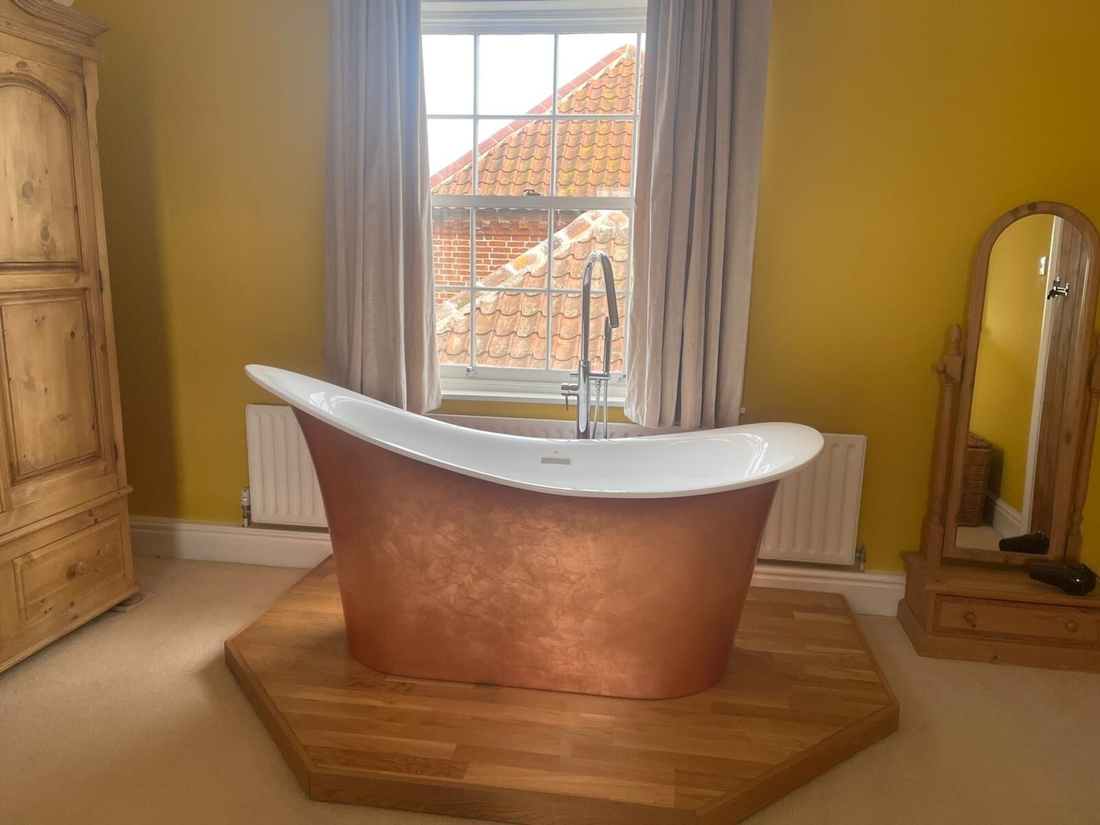 Un baño con una bañera independiente, un armario de madera, una ventana con cortinas y un espejo.