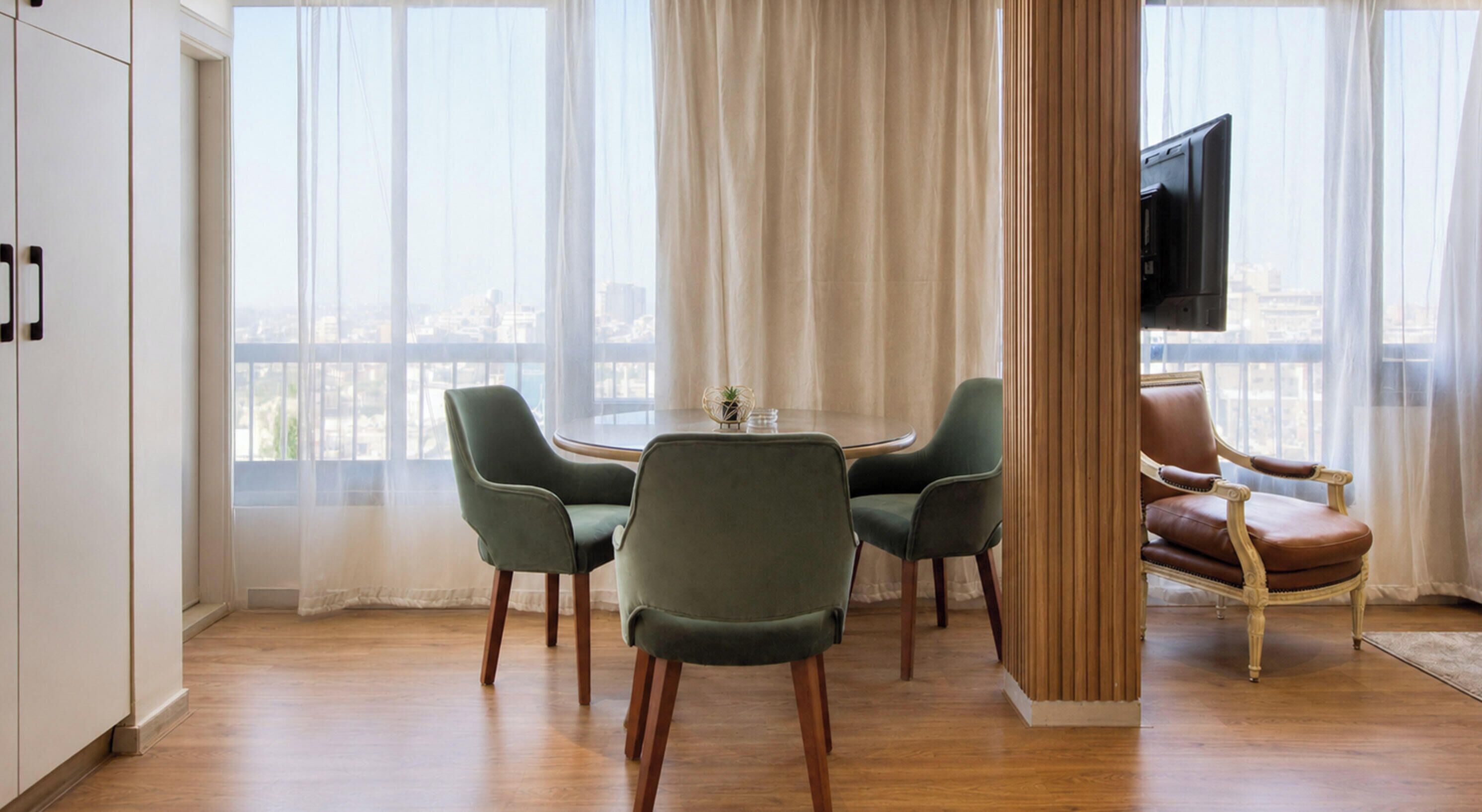 Uma sala de jantar com uma mesa redonda e quatro cadeiras, uma televisão na parede e vista para a cidade através de grandes janelas.
