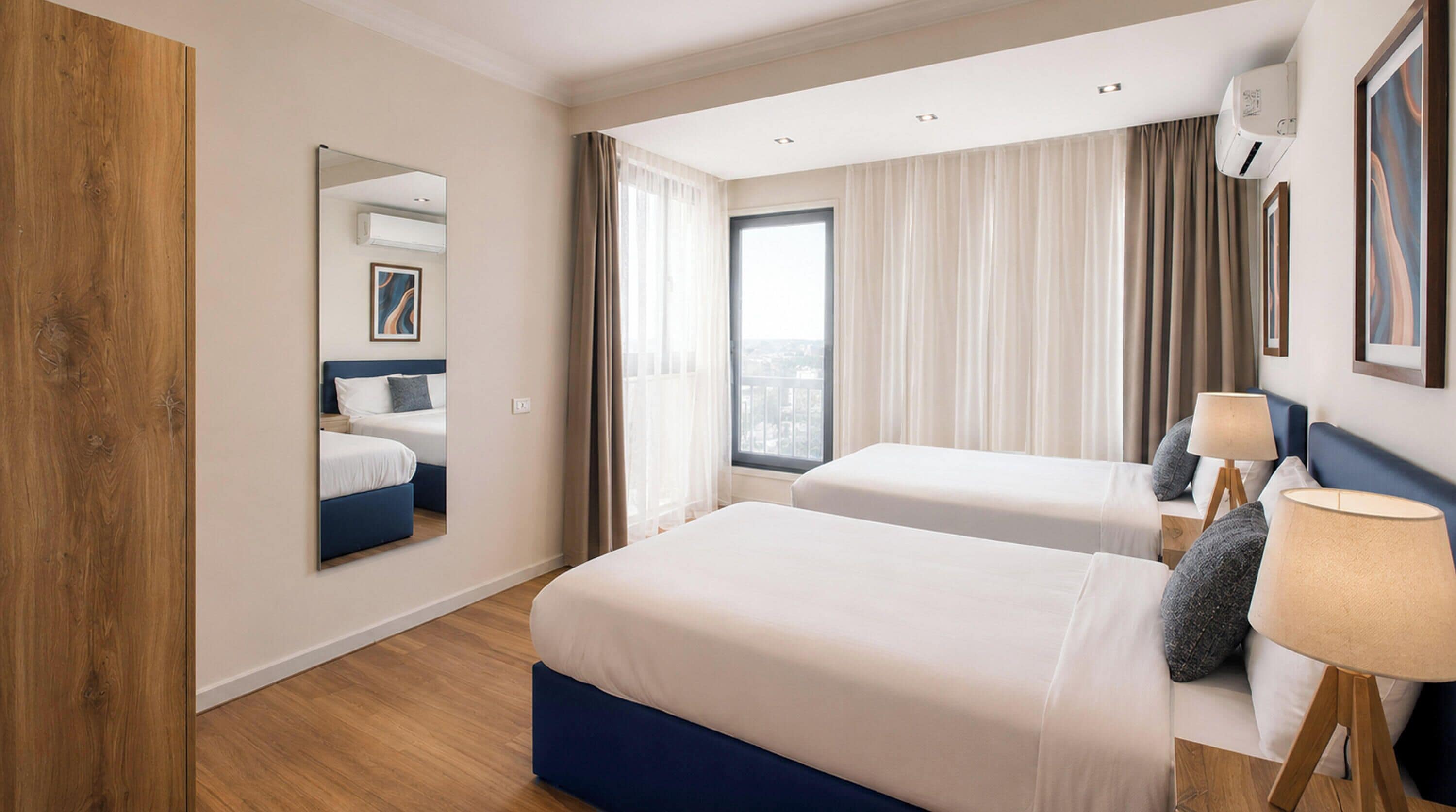 Une chambre d’hôtel avec deux lits, une grande fenêtre, un miroir et un climatiseur fixé au plafond.