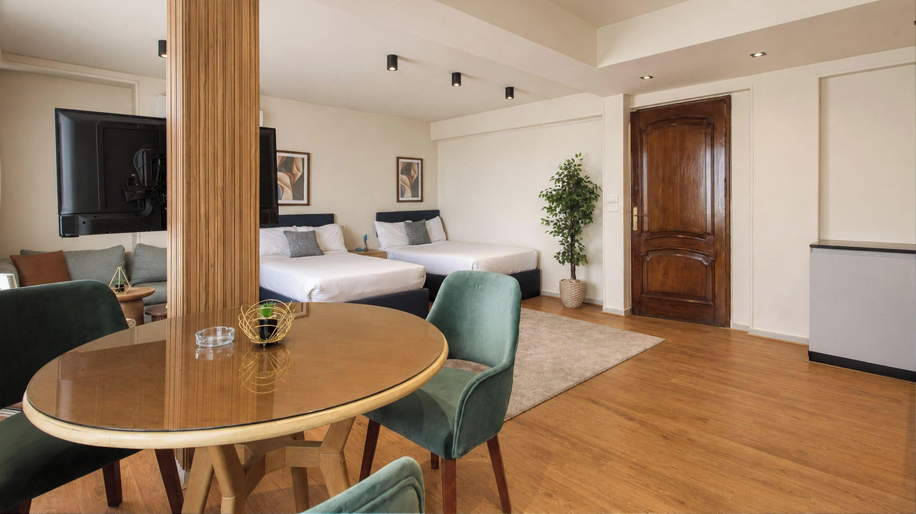 A modern hotel room with a round glass table, two single beds, a wooden door, and a small potted plant.