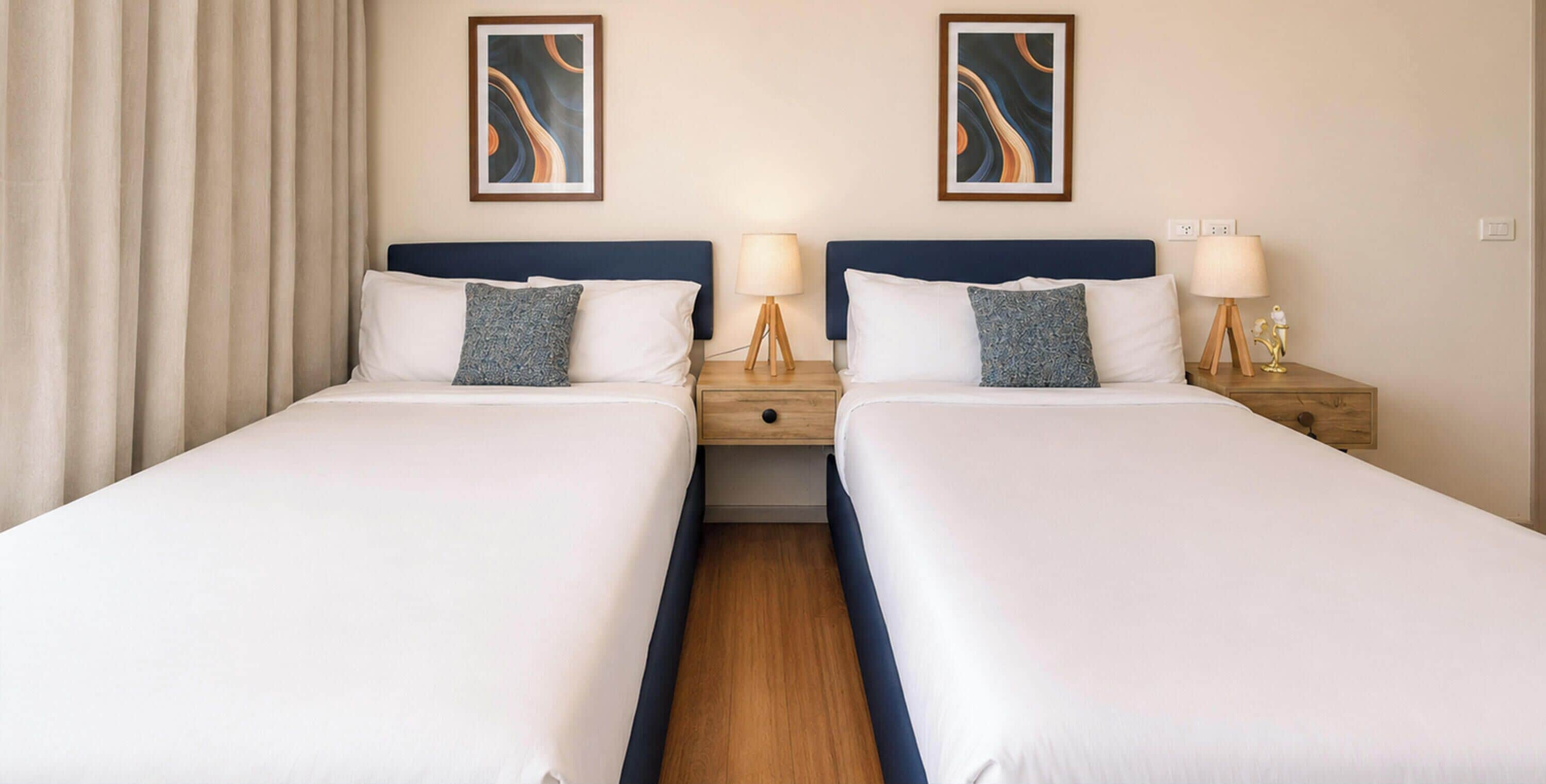 A hotel room with two beds, each with a headboard, bedside tables, lamps, and a curtain on the left side.