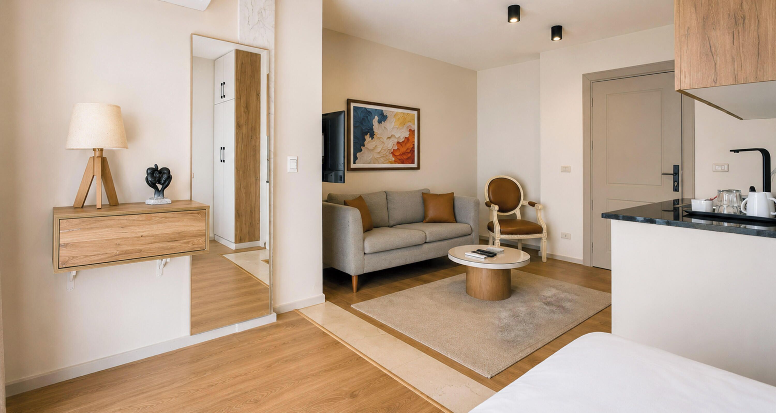 A modern hotel room with a wooden floor, a sofa, a round coffee table, a chair, a painting on the wall, and a kitchenette.