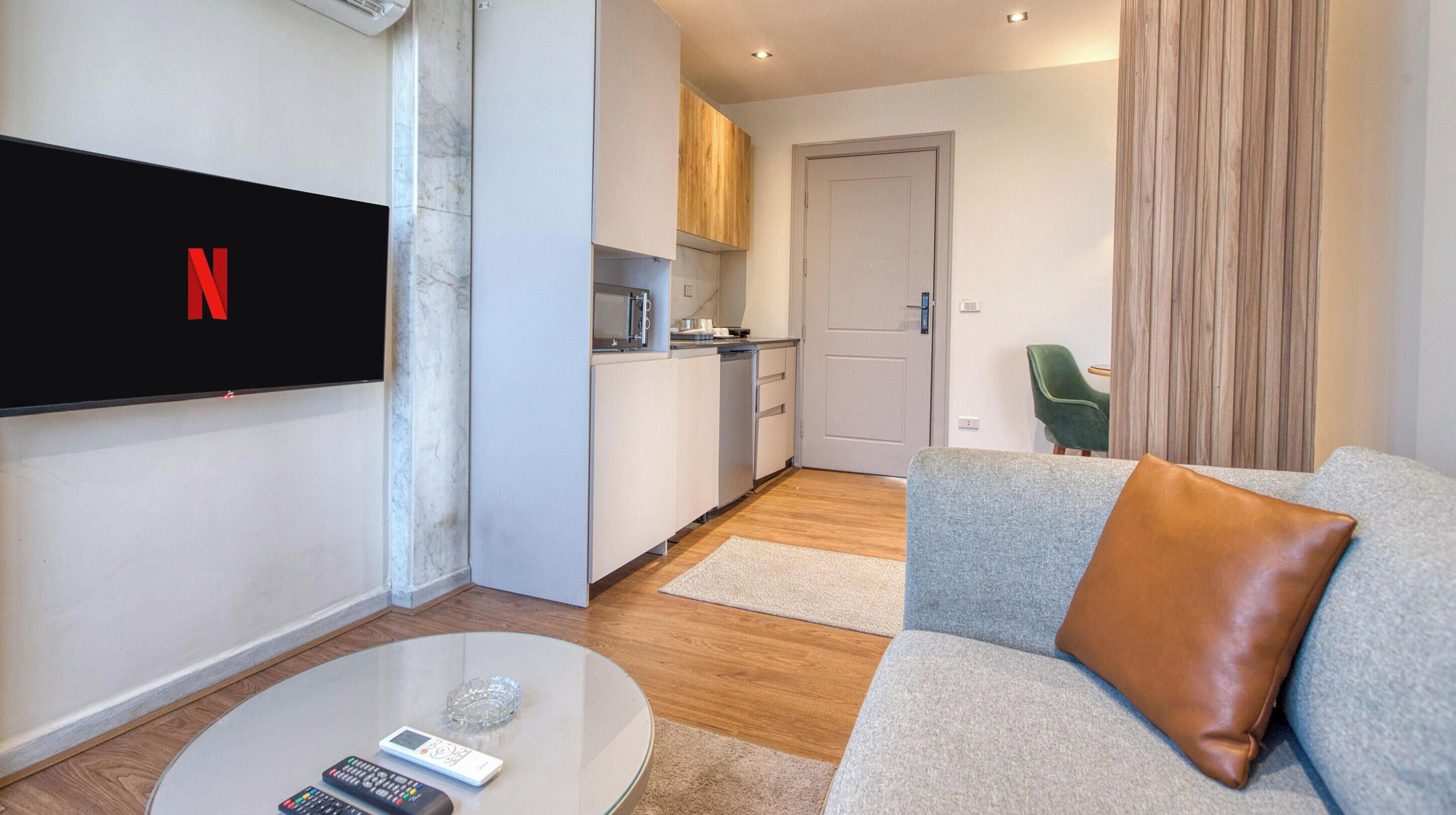 A modern living room with a flat-screen TV, a coffee table with remote controls, a grey sofa with a brown cushion, and a kitchen area with wooden cabinets and a microwave.