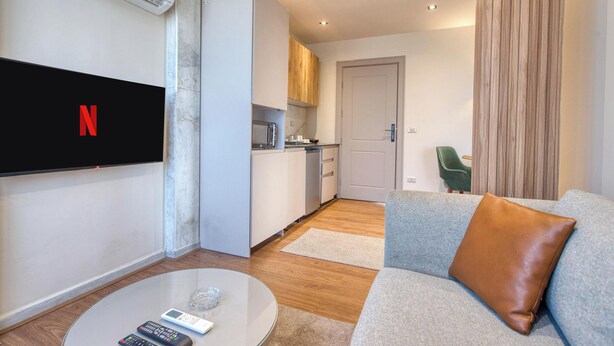 Una sala de estar moderna con un televisor de pantalla plana, una mesa de centro con controles remotos, un sofá gris con un cojín marrón, y una zona de cocina con armarios de madera y un microondas.