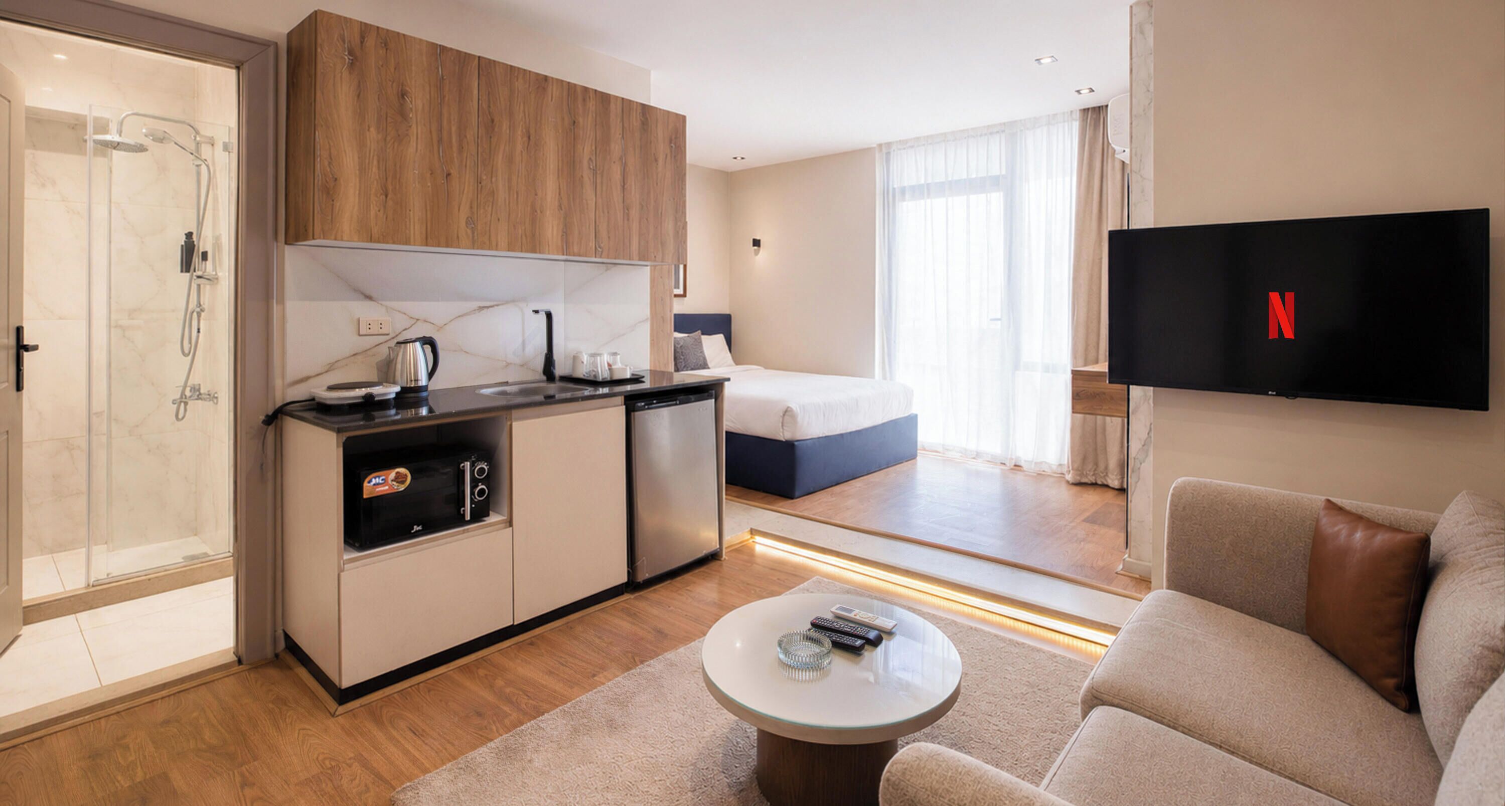A modern hotel room with a kitchenette, a bed, a TV, and a seating area.