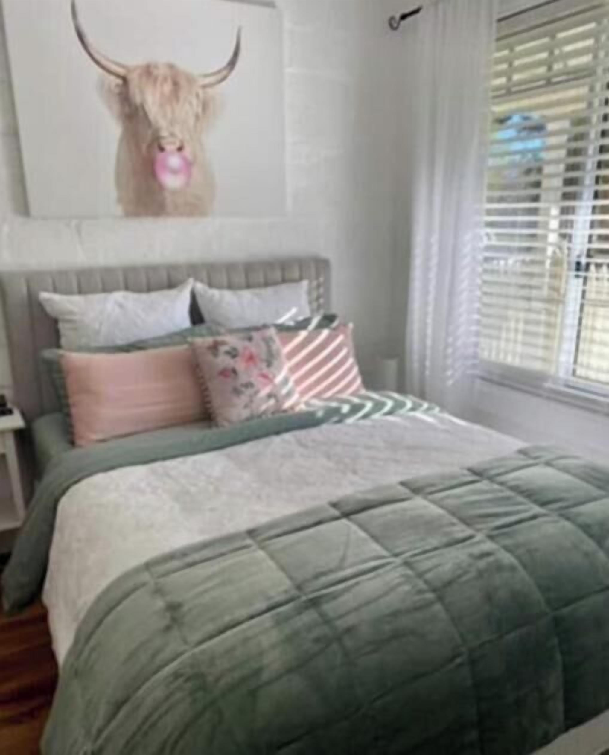 Un lit recouvert d’une couette verte, une tête de lit ornée d’une décoration représentant une tête de taureau et une fenêtre avec des stores.