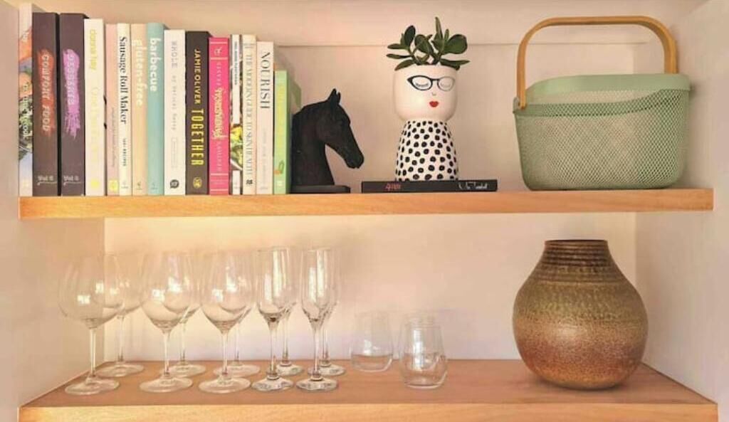 Uma estante com livros, uma estatueta de cavalo, uma planta em vaso, uma cesta verde e copos de vinho.