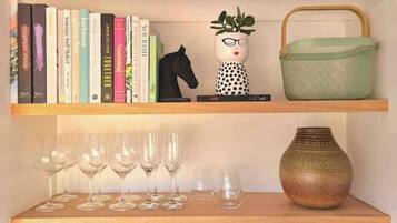 Uma estante com livros, uma estatueta de cavalo, uma planta em vaso, uma cesta verde e copos de vinho.