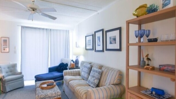 Ein Wohnzimmer mit Sofa, Sessel und Couchtisch. Ein Bücherregal mit Dekorationselementen und ein Deckenventilator sind ebenfalls vorhanden.