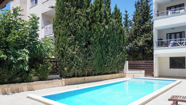 A rectangular swimming pool with a tiled edge, surrounded by a concrete deck. There are wooden lounge chairs and a wooden fence with a gate. - Spacious apartamens nearthe mall (Pafos)