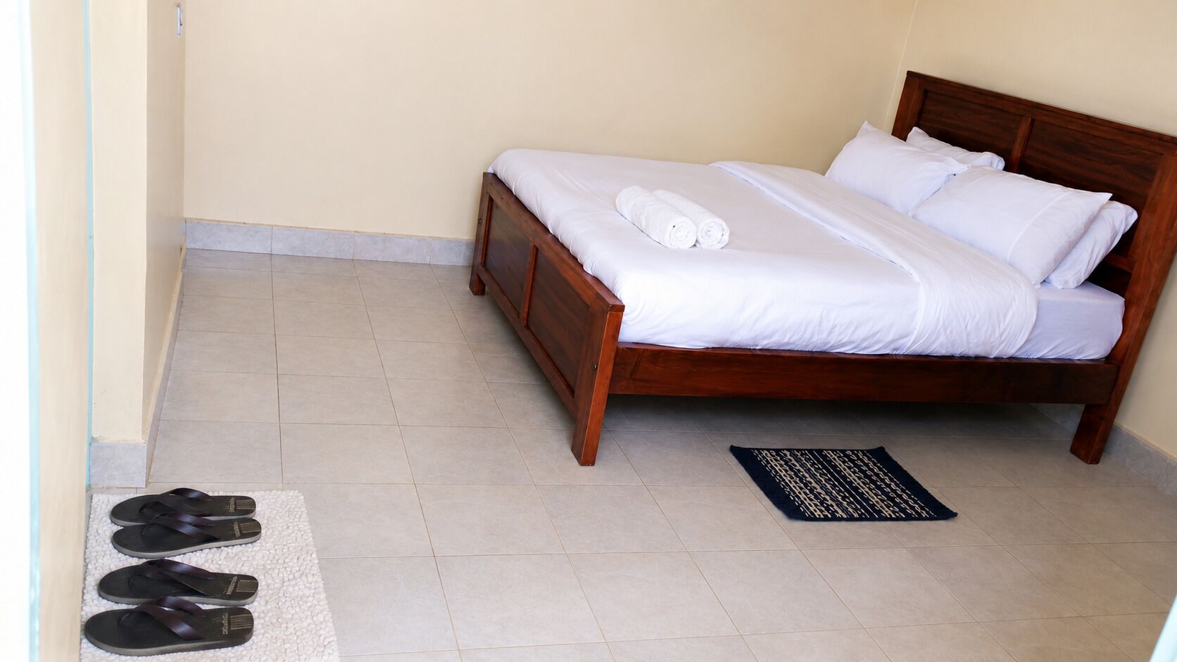 A hotel room with a bed, a pair of slippers, and a doormat.