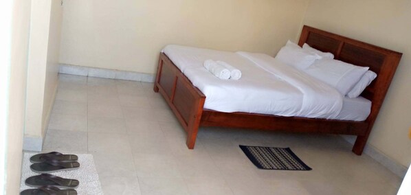 A single bed with white bedding and two rolled towels, a wooden headboard, and a small rug on a tiled floor.