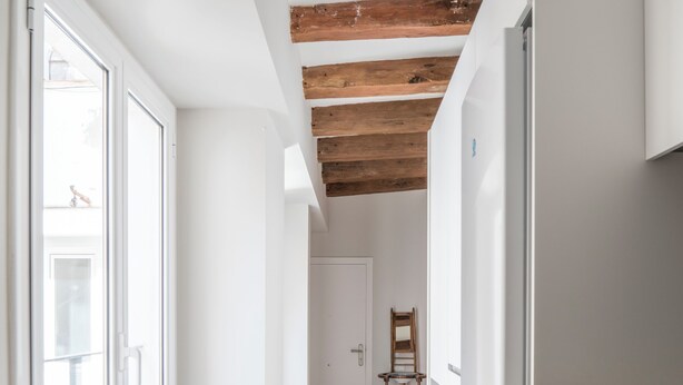 Ein schmaler Flur mit einer Küche an der Seite, Holzbalken an der Decke und einem Stuhl in der Ferne.
