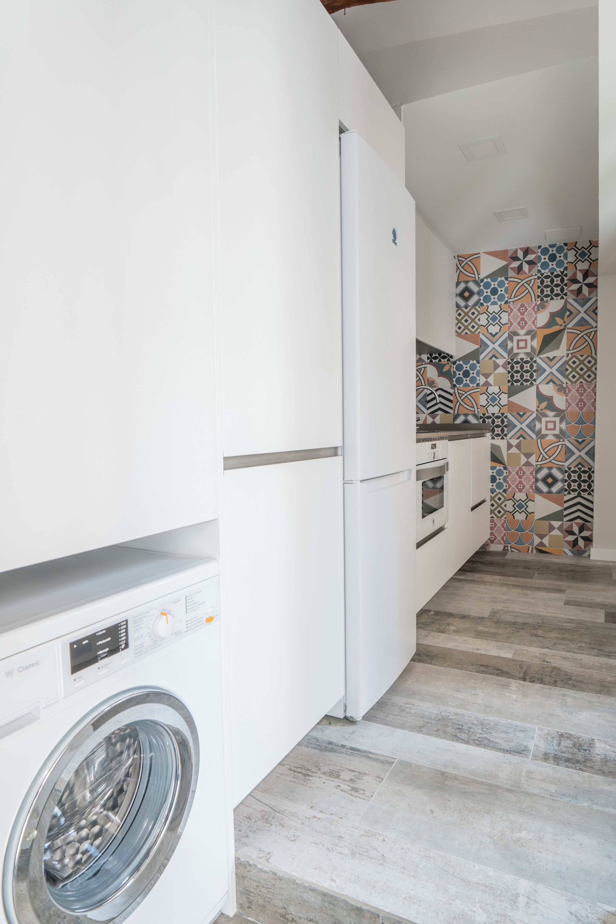 A compact kitchen with a washing machine, a refrigerator, and a patterned backsplash.
