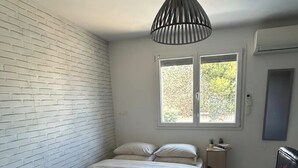 Quarto com parede de tijolos brancos, cama com roupa de cama branca, luminária pendente, janela com vista e uma pequena mesa de cabeceira.