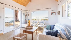 Um quarto amplo com teto alto, uma grande janela com vista para a praia, uma mesa de madeira e um sofá com almofadas listradas.