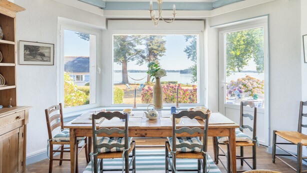 Uma sala de jantar com mesa, cadeiras, um lustre e vista para o exterior.