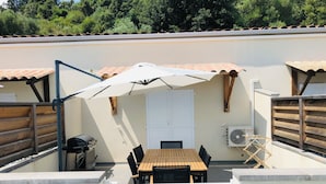 En udendørs patio med et træbord, stole, grill og en hvid parasol.
