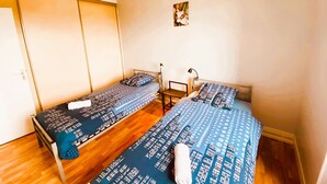 Quarto com duas camas, soalho de madeira, luminária no teto e uma pequena mesa de cabeceira com abajur.