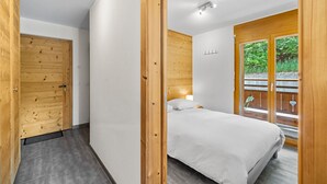 A bedroom with a wooden door, a bed with white bedding, and a view of greenery through a window. - Sandra's Farm F0 - Cozy with a Terrace, in the Center (Haute-Nendaz)