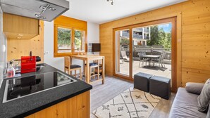 A modern kitchen with a black countertop, wooden cabinets, and a red appliance. There is a dining area with a table and chairs, and a large sliding glass door leading to an outdoor patio. - Sandra's Farm F0 - Cozy with a Terrace, in the Center (Haute-Nendaz)