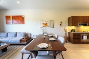 A modern living space with a dining table set for four, a sofa, a kitchen with a microwave, and a large abstract painting on the wall. - Chic 2BR | Beach, Town & Elevated Views (Huatulco)
