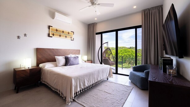 Ein Schlafzimmer mit Bett, Nachttischen, Schreibtisch, Stuhl, Deckenventilator, großem Fenster und Blick ins Grüne.
