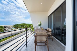 A balcony with a dining table and chairs, a vase with flowers, and a view of a road and greenery. - Chic 2BR | Beach, Town & Elevated Views (Huatulco)