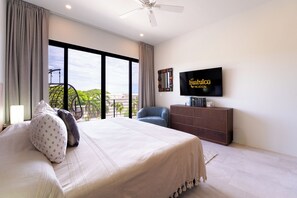 A hotel room with a bed, a TV, a chair, a balcony, and a view of the city. - Chic 2BR | Beach, Town & Elevated Views (Huatulco)