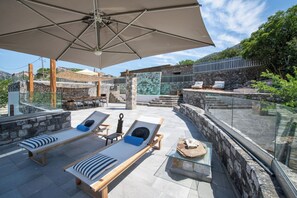 Eine Dachterrasse mit Liegestühlen, einem Tisch und einer Glasbrüstung mit Blick auf eine gebirgige Landschaft.
