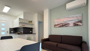 A modern kitchen with white cabinets, a dining table with chairs, a brown sofa, and a beach scene painting on the wall. - Beachfront, Modern Flat With Lovely Garden (San Michele al Tagliamento)