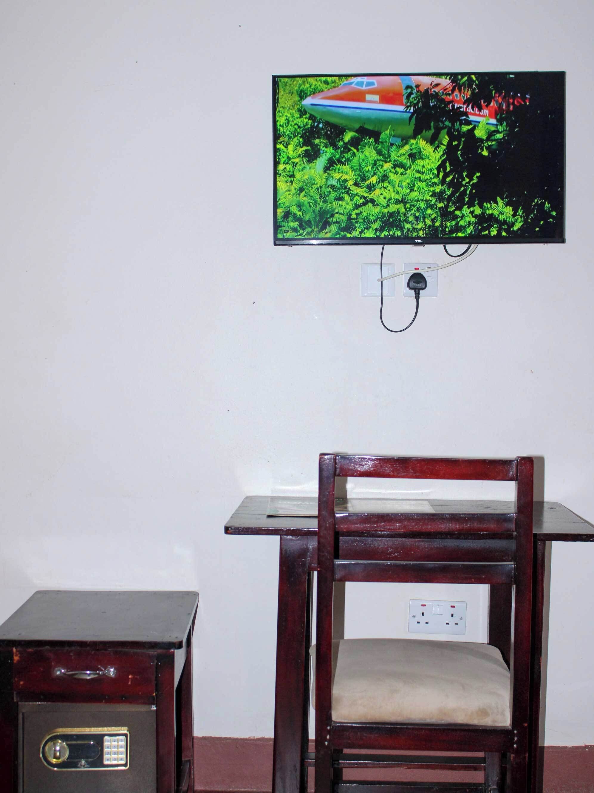 Quarto com uma cadeira de madeira, uma mesinha, um cofre e uma televisão fixada na parede.
