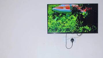 Quarto com uma cadeira de madeira, uma mesinha, um cofre e uma televisão fixada na parede.