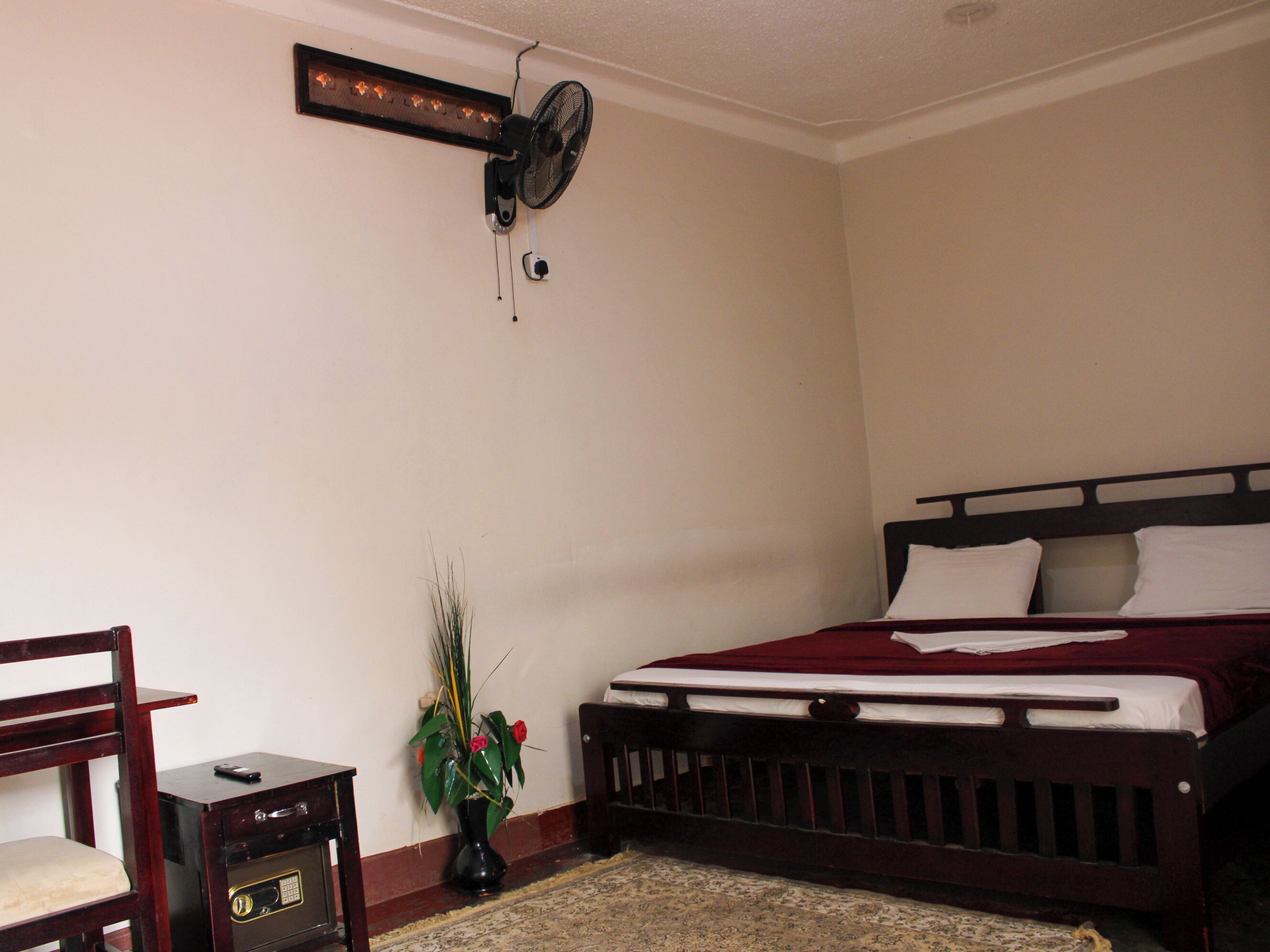 A bedroom with a bed, a chair, a small table, a wall-mounted fan, and a potted plant.