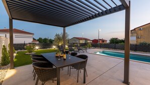 Een overdekte patio buiten met een zwembad, eettafel en stoelen.