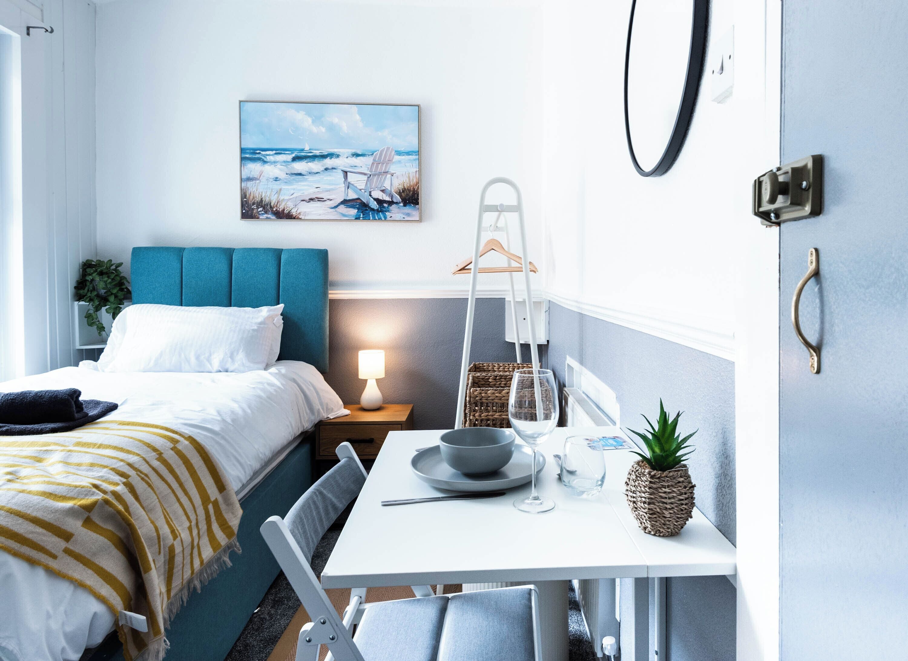 A hotel room with a bed, a table with a bowl and wine glass, a chair, a hanging plant, and a painting of a beach scene.