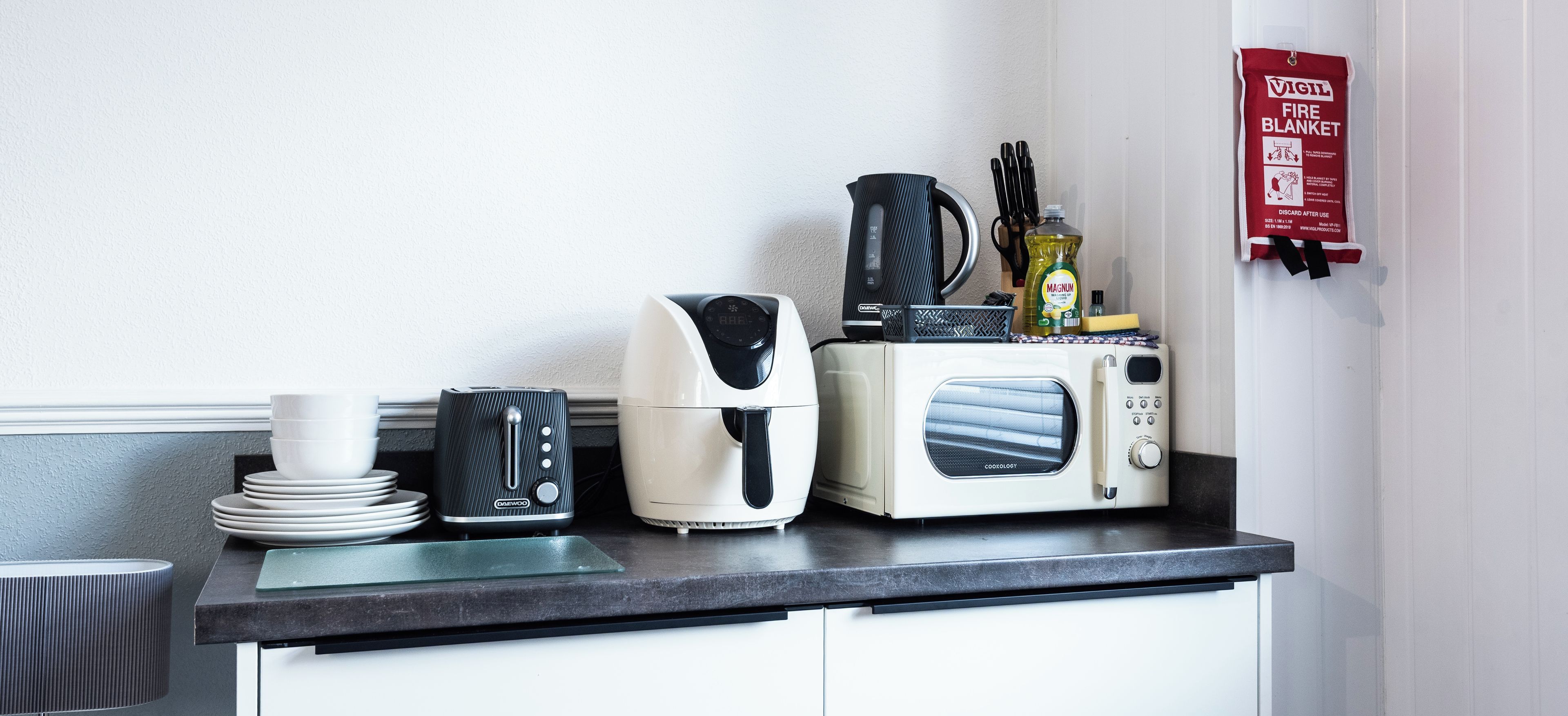 Una encimera de cocina con cafetera, tostadora, hervidor y microondas. Hay una manta anti-incendios colgada en la pared.